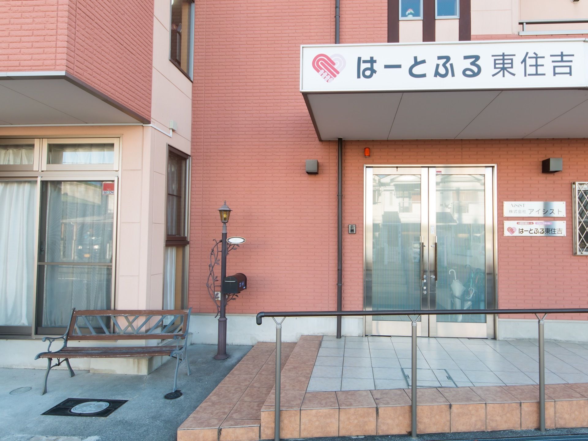 ピンク色の外壁が施された建物。施設名称が明記された屋根の下に扉があり、手前に手すりがある。
