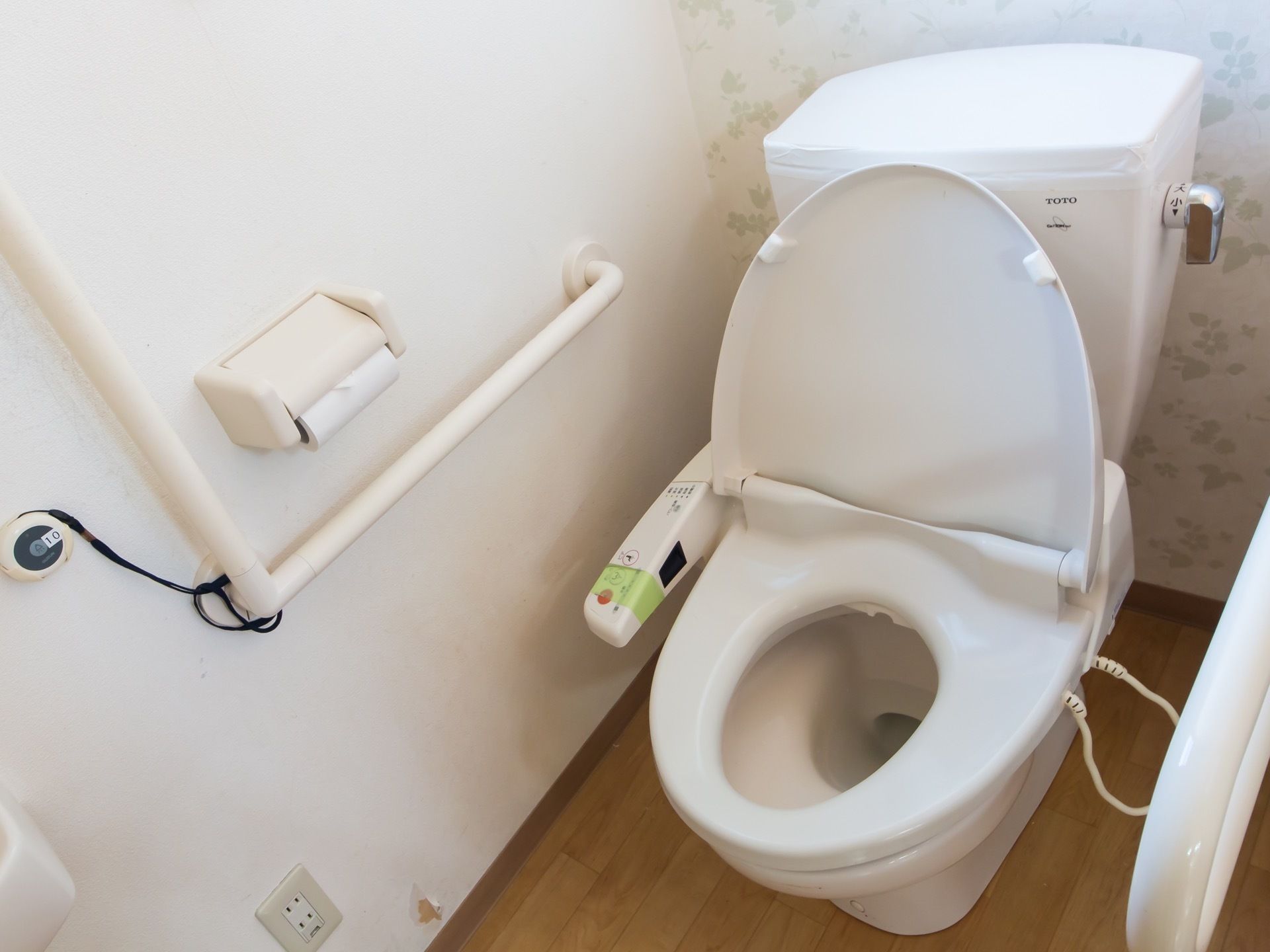花柄の壁紙が施されたトイレスペースに洋式便器がある。両側の壁面に白い手すりが取り付けてある。