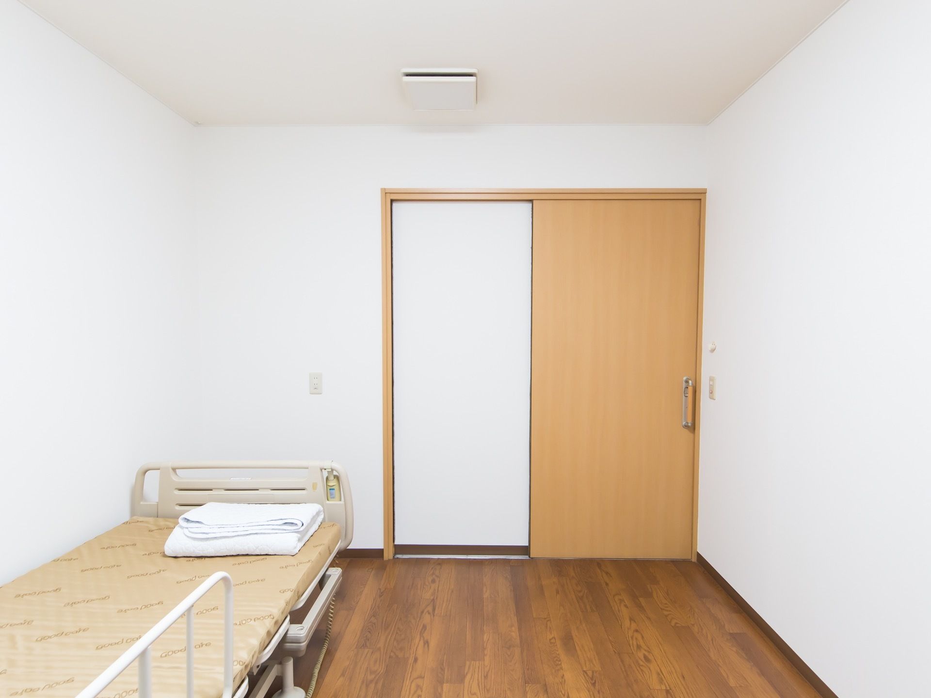 木目が美しいフローリングの室内にベッドが据えてある。天井や壁面が白く、木目調の引き戸がある。