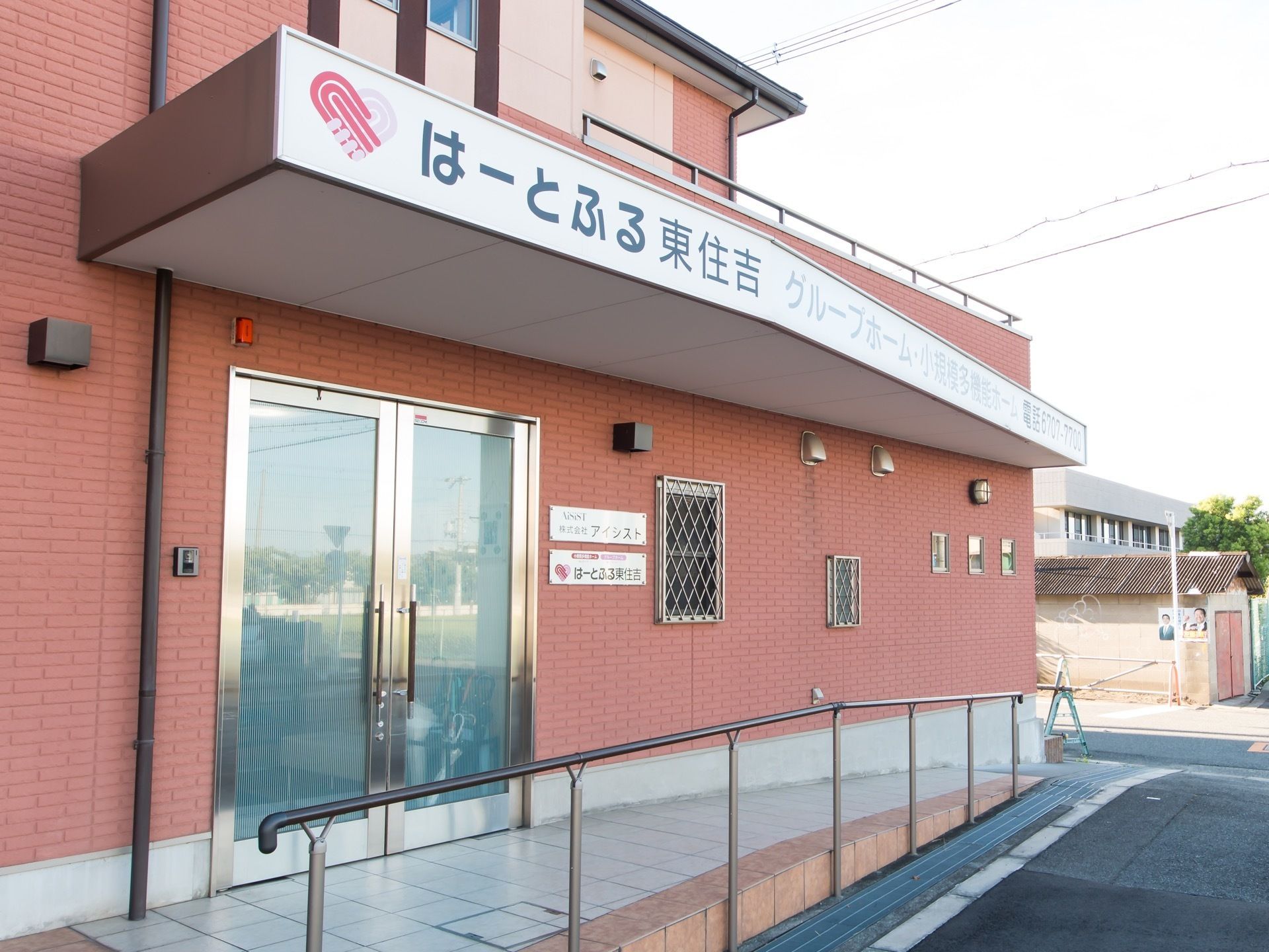 舗装された敷地にピンク色の建物がある。施設名称が記された屋根の下にスロープと手すりがある。