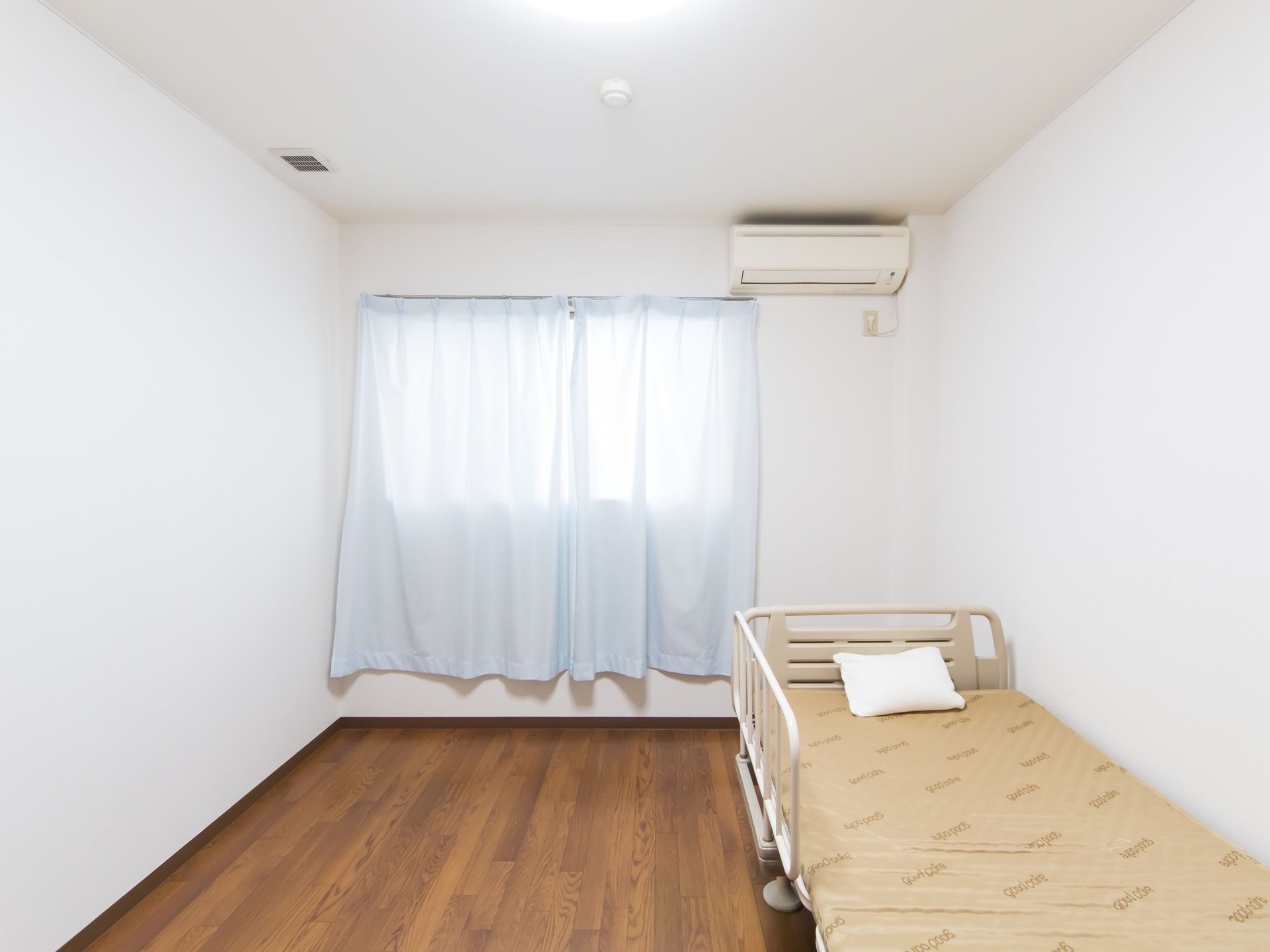 木目が美しい茶色のフローリングの室内にベッドがある。窓には長めの淡い水色のカーテンが取り付けてある。