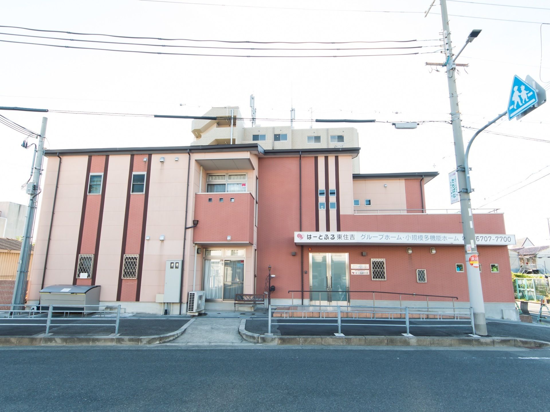 車道に面して、2階建てのピンク色の施設が建っている。建物の前にはアスファルトで舗装された歩道がある。