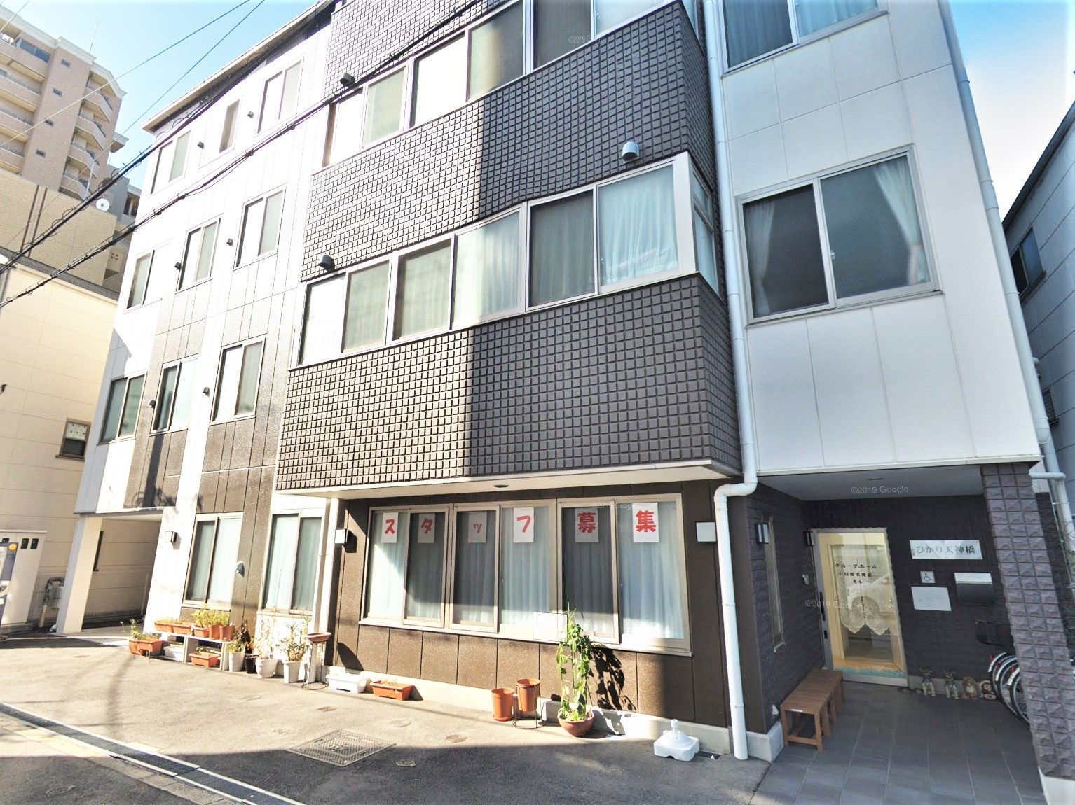 日当たりのいい４階建ての建物の周辺にはプランターがたくさん置かれ、玄関前にはベンチもある。
