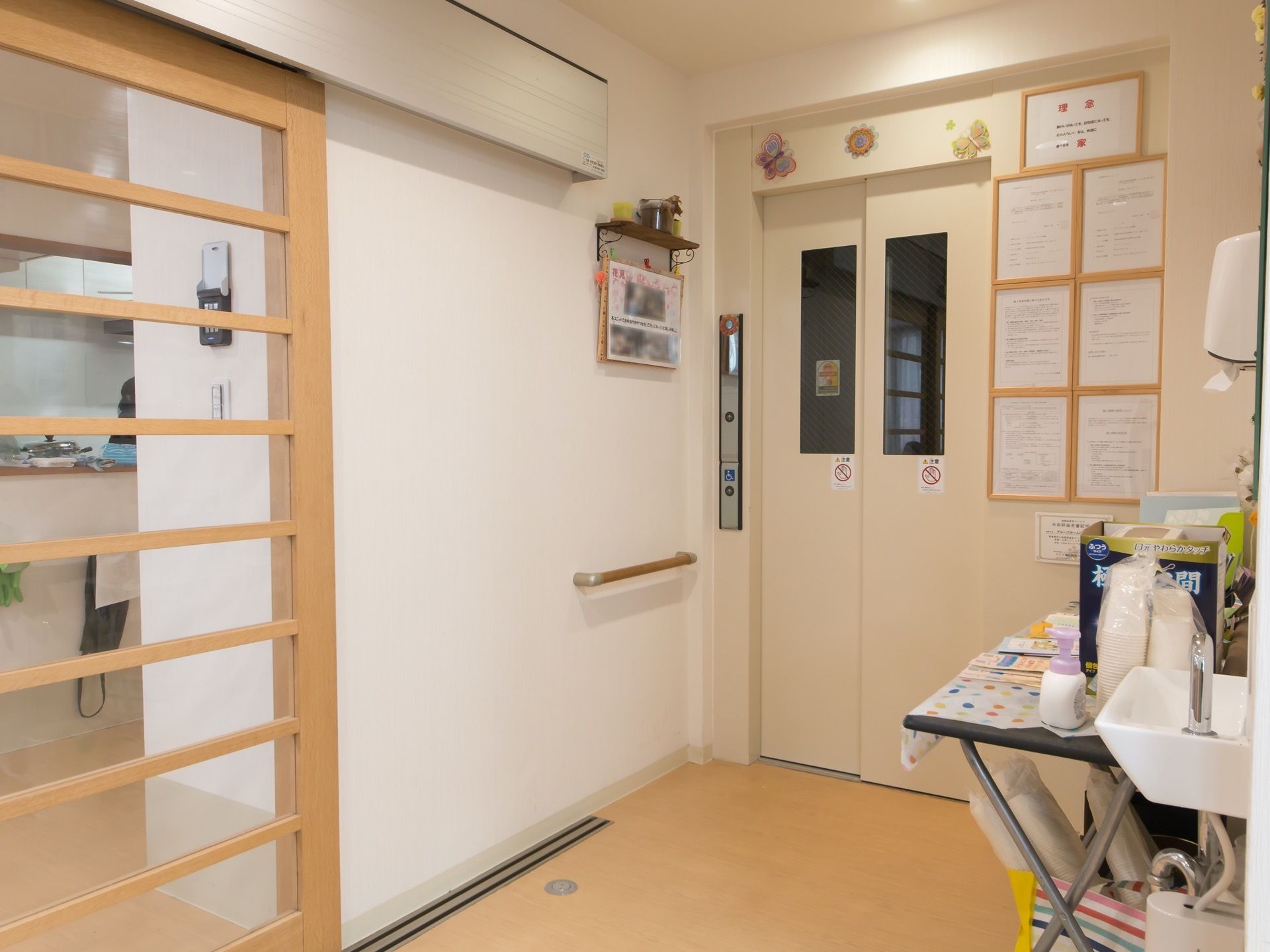 白を基調とした扉のエレベーターが１基設置されている。手前には手洗い場が設けられており、横の机には紙コップやハンドソープが用意されている。