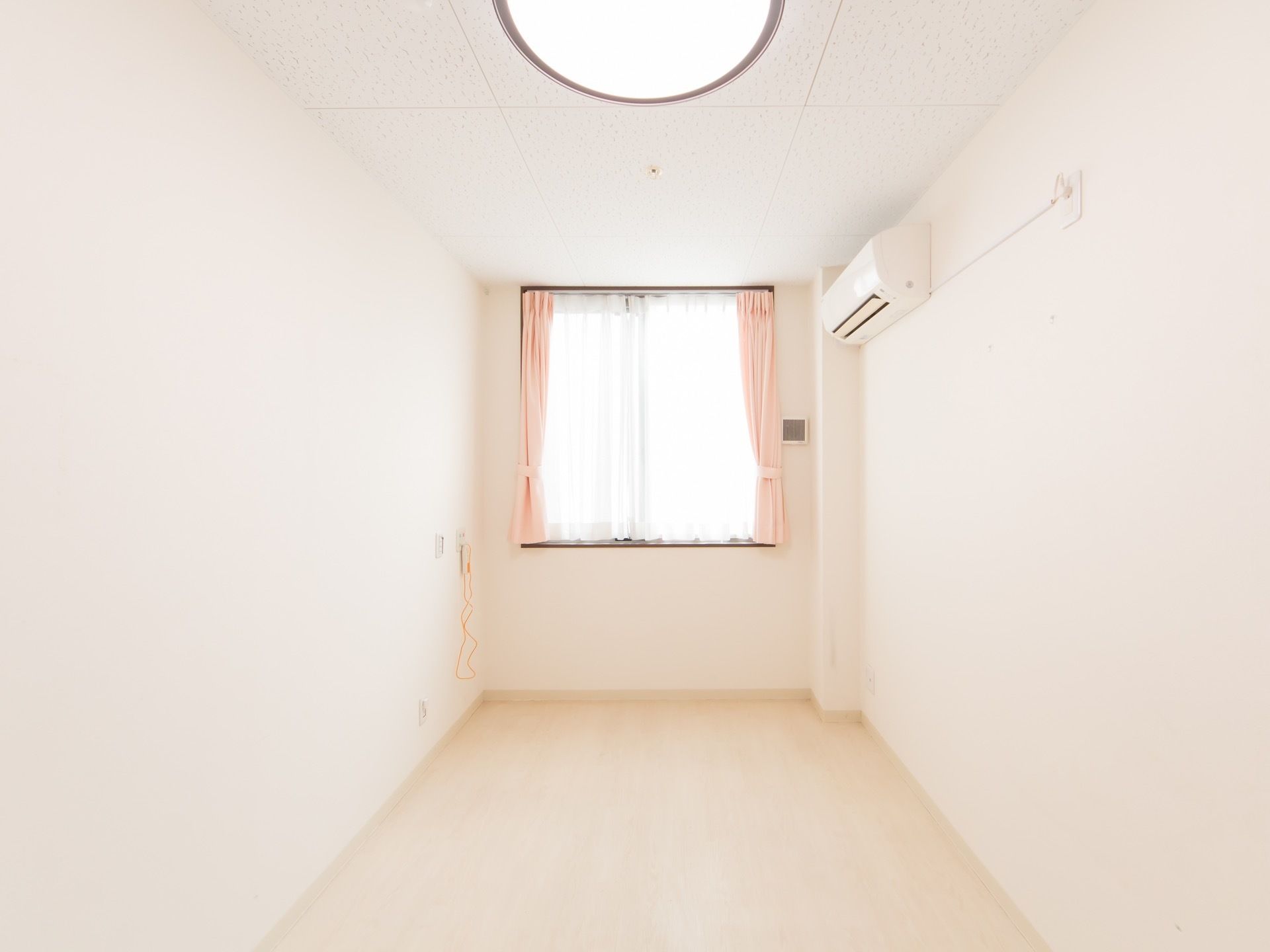 白を基調としたワンルームの居室。天井には照明が設置されており、窓にはレースとカーテンが取り付けられている。