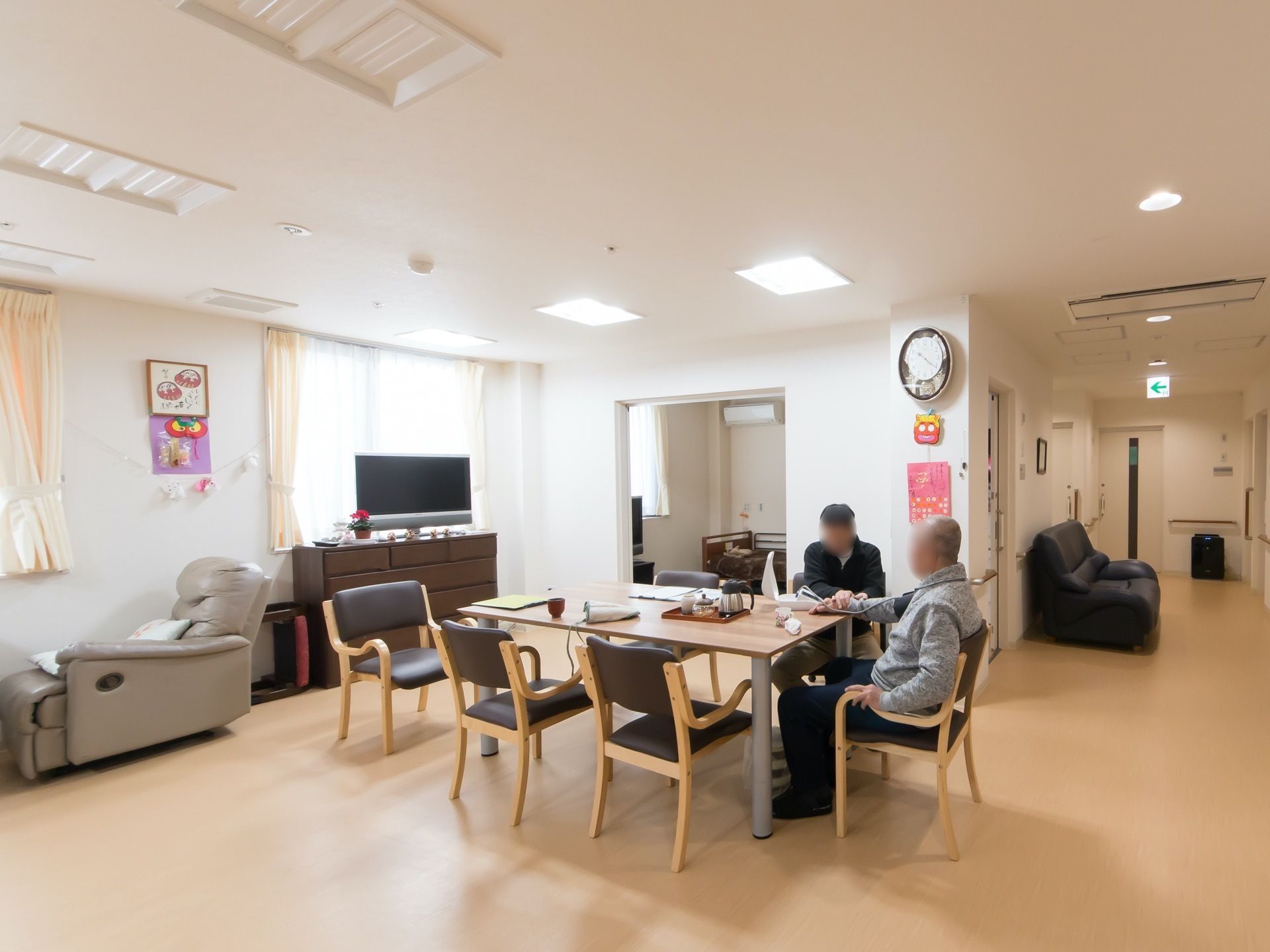 明るく開放的な食堂で、椅子に座って入居者様が寛いでいる。窓際には、テレビやマッサージチェアが置かれている。