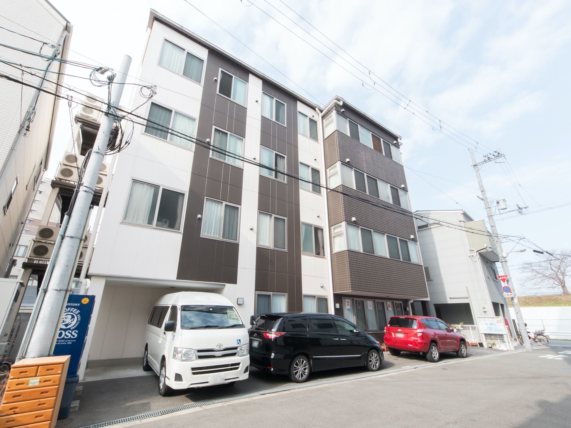白色と茶色を基調とした、近代的なデザインのマンションのような４階建ての建物である。道路沿いのスペースに車が３台停められている。