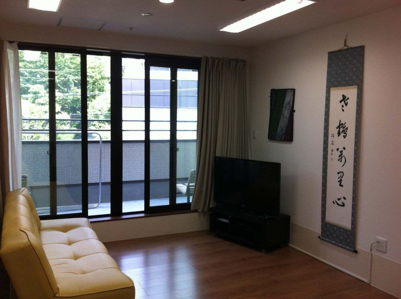 ベランダ付きのリビング。リビングにはテレビやソファがあり、掛け軸が飾られている。ベランダにはいすが置かれている。