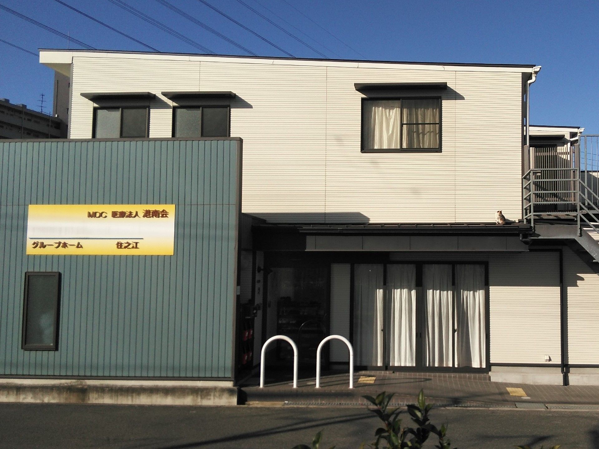 建物を正面から見た画像。2階建ての白を基調とした建物で、手前のモスグリーンの外壁にホームの看板がある。
