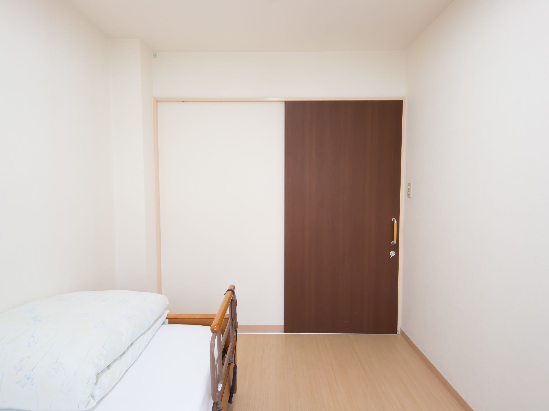 白い壁に木目調の床、ブラウンの扉が設置されたシンプルな居室である。手前にはベッドが置かれ、床には障害物がなくスッキリとしている。