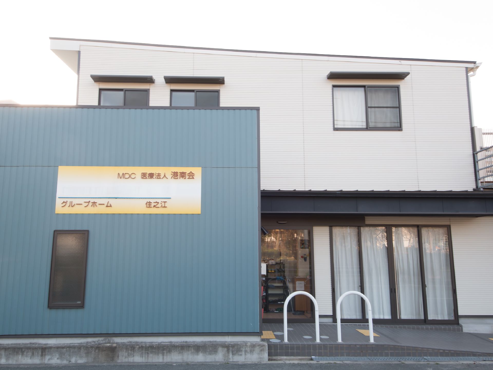 白とブルーグレー調の２階建て施設建物である。淡い黄色のプレートに施設名が書かれている。入口手前には、スロープと手すりが設置されている。