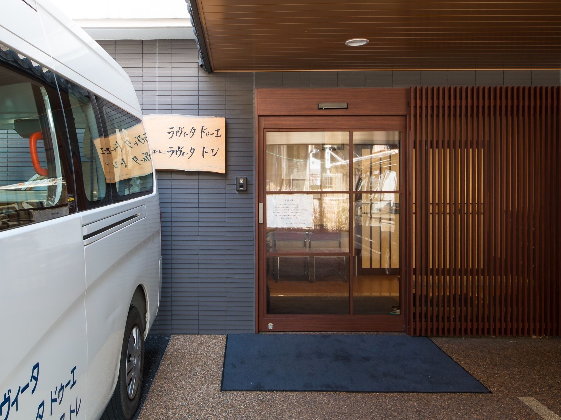 ガラス張りの玄関ドアがある。周囲は木製の格子で覆われている。壁は青く、玄関ドアの上はルーフが設置されている。