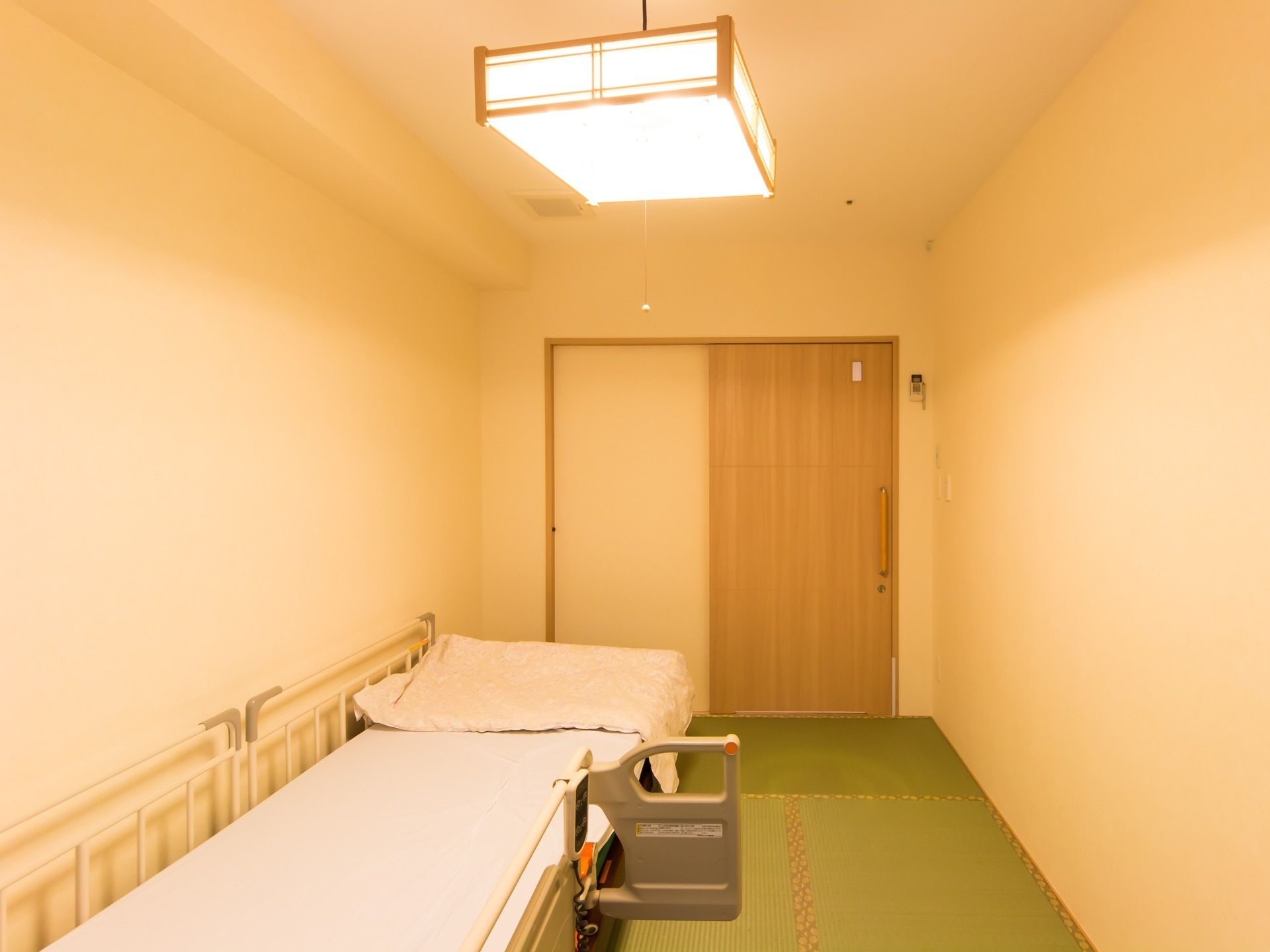 木目調の引き戸が付いた居室がある。床には畳が敷かれ、和風のペンダントライトが下がっている。介護用ベッドがある。