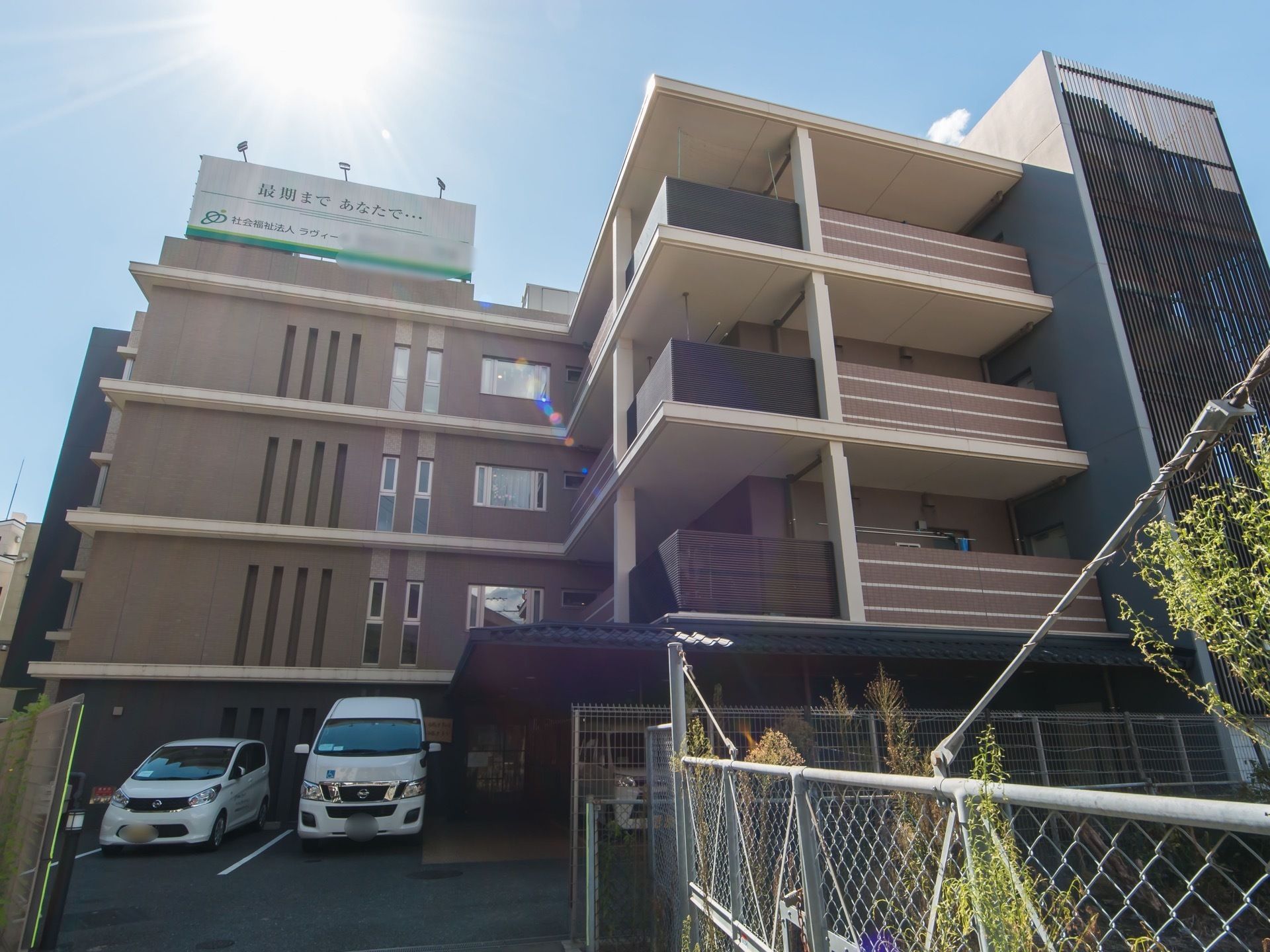 茶色い壁の４階建ての大きな建物がある。敷地内には駐車場がある。建物の屋上部分には大きな看板が立っている。