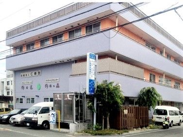 グレーとオレンジの建物は3階建てで、ベランダや駐車場も設置され、周辺には住宅がある。