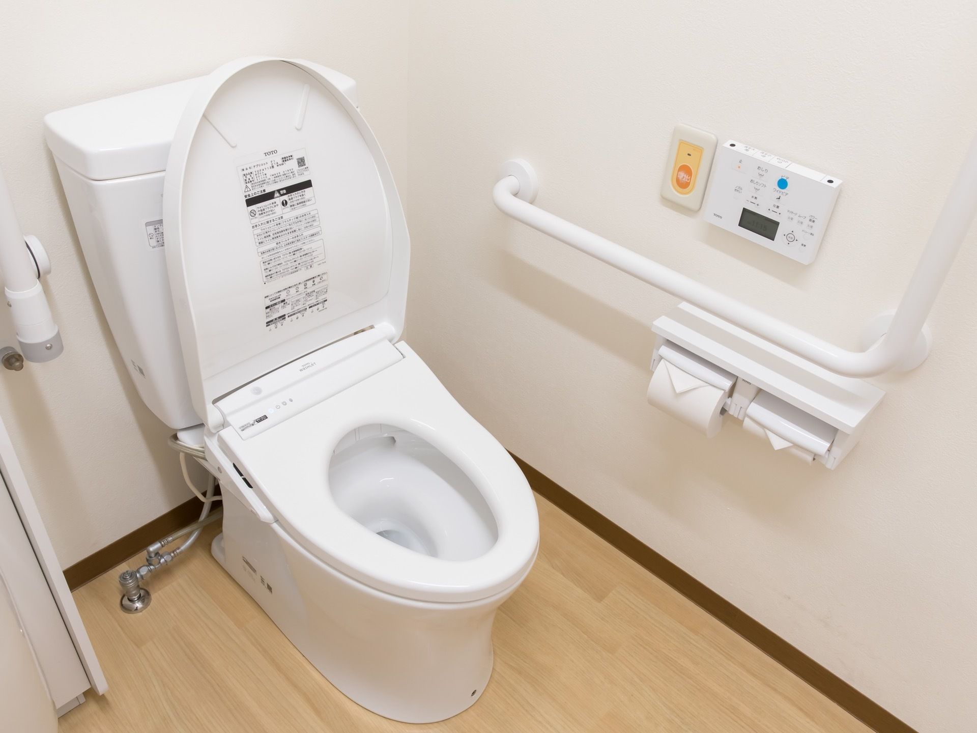 トイレには、温水洗浄便座が完備されている。白い壁には操作パネルや緊急呼び出しブザー、L字型手すりが設置されている。