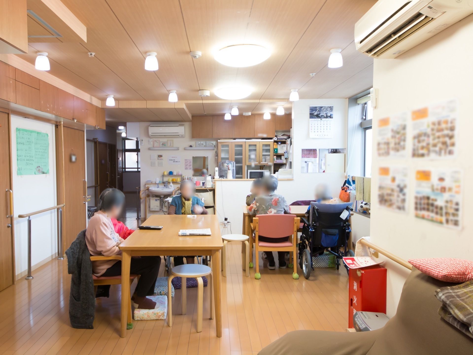 食堂の天井には照明器具がたくさんついている。椅子の前には小さな台があり、入居者様がそこに足を置いて座っている。