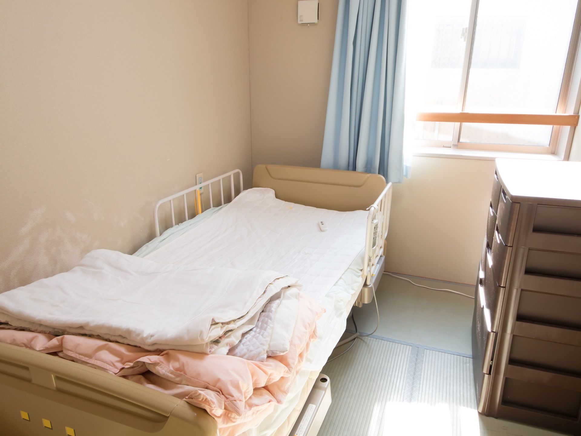 畳敷きの居室に、転落防止柵つきの介護ベッドと衣類収納タンスが置かれている。窓には、木の手すりがついている。