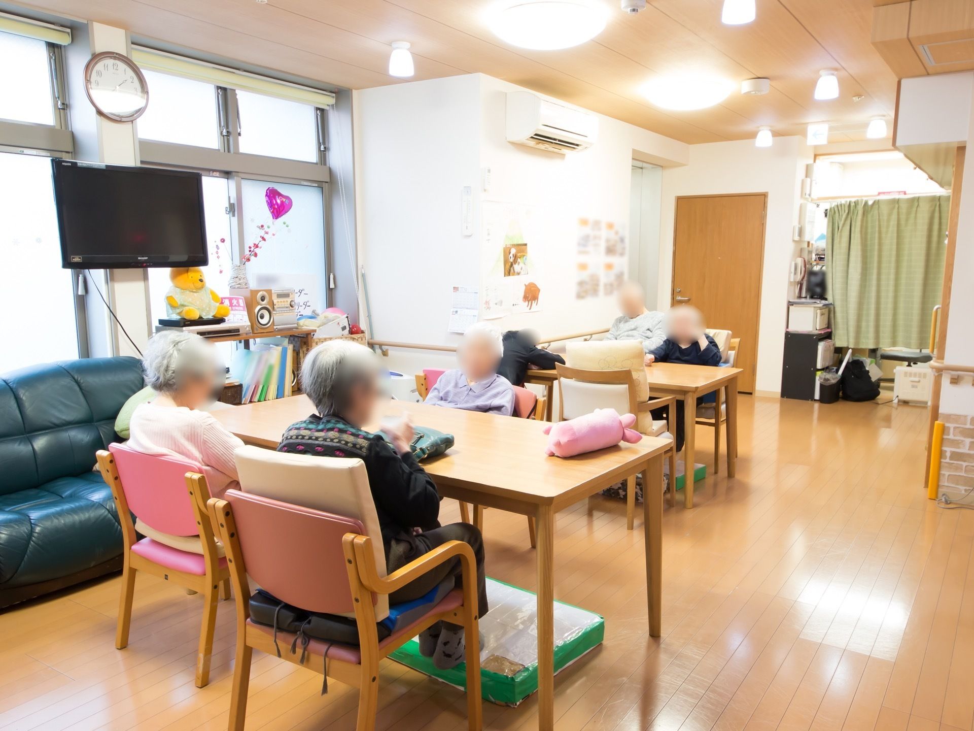 共有スペースに入居者様が６名座っている。テーブルの下には、足置き台が置かれている。照明器具が多くついている。