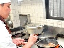 白いコック帽とコック服を身につけた料理人がフライパンで炒め物をしている様子。