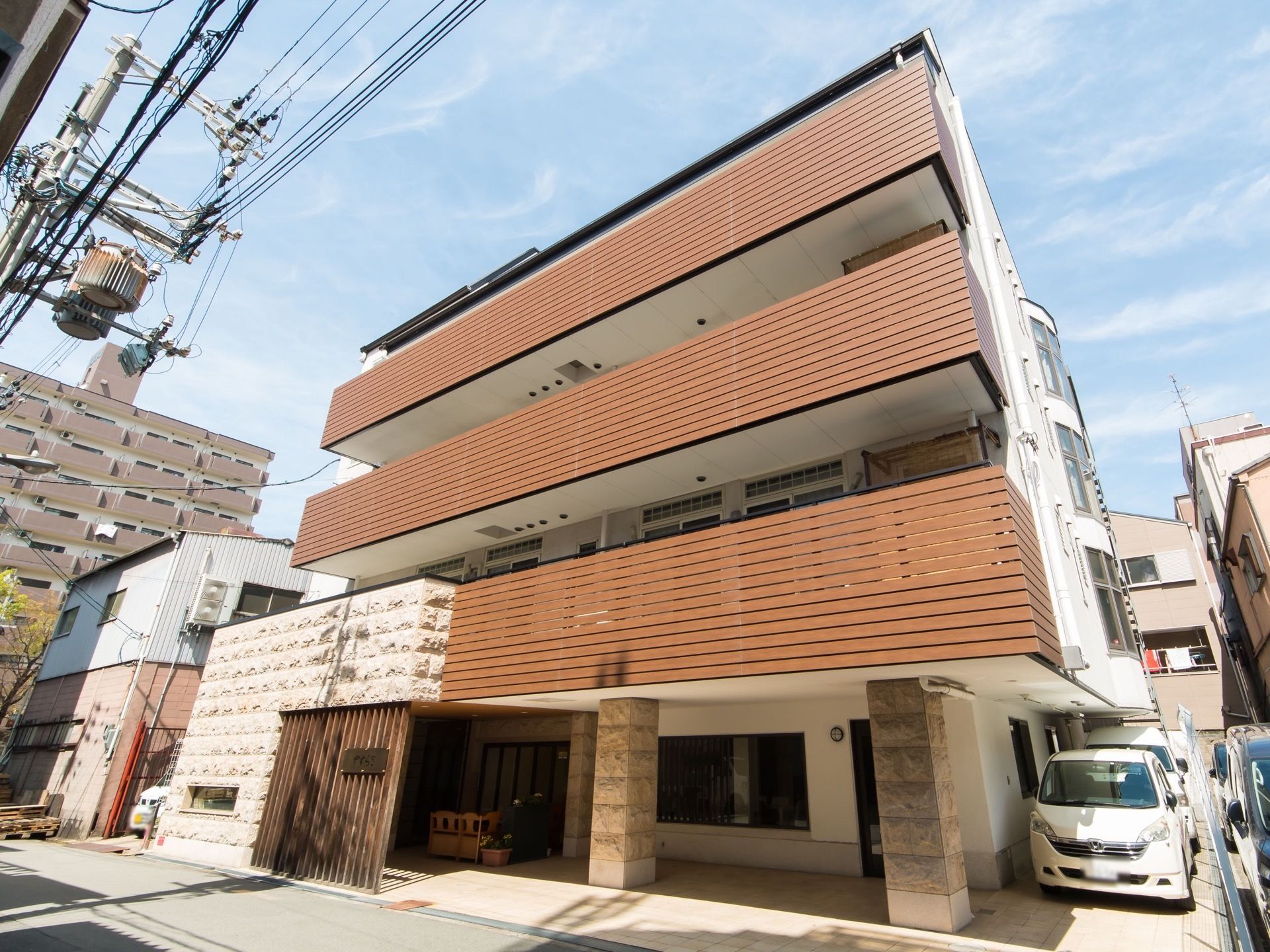 ３階建てのデザイン性ある外観の施設である。風通しの良い場所にテーブルセットが置かれ、駐車スペースを完備している。