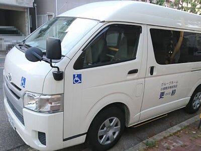 白いバン型の大きな福祉車両。白い車体にはブルーの文字でホーム名が書かれていて、青い車いすマークのステッカーも複数個所に貼られている。