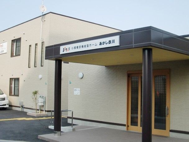 ベージュの2階建ての建物。駐車場があり、玄関には手すり付きのスロープや屋根が付いている。