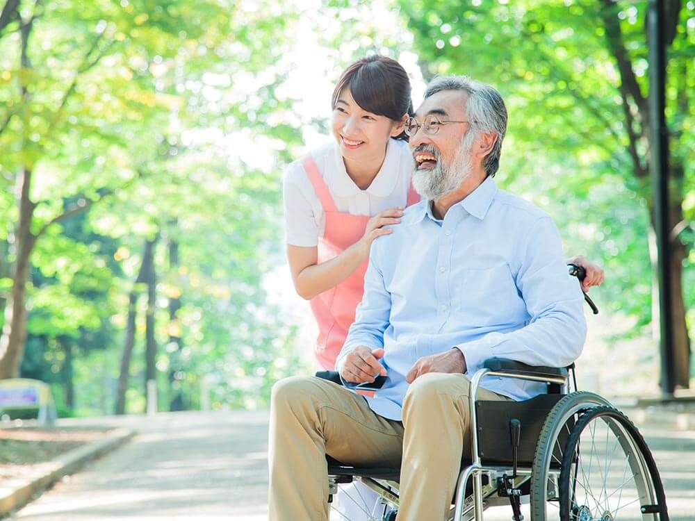 木陰にいる車椅子に乗った高齢男性と介助をする女性ヘルパー