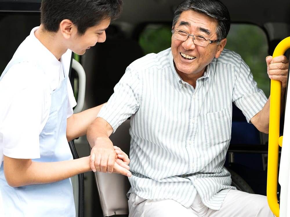 手すりにつかまり車に乗る高齢男性を介助するヘルパー