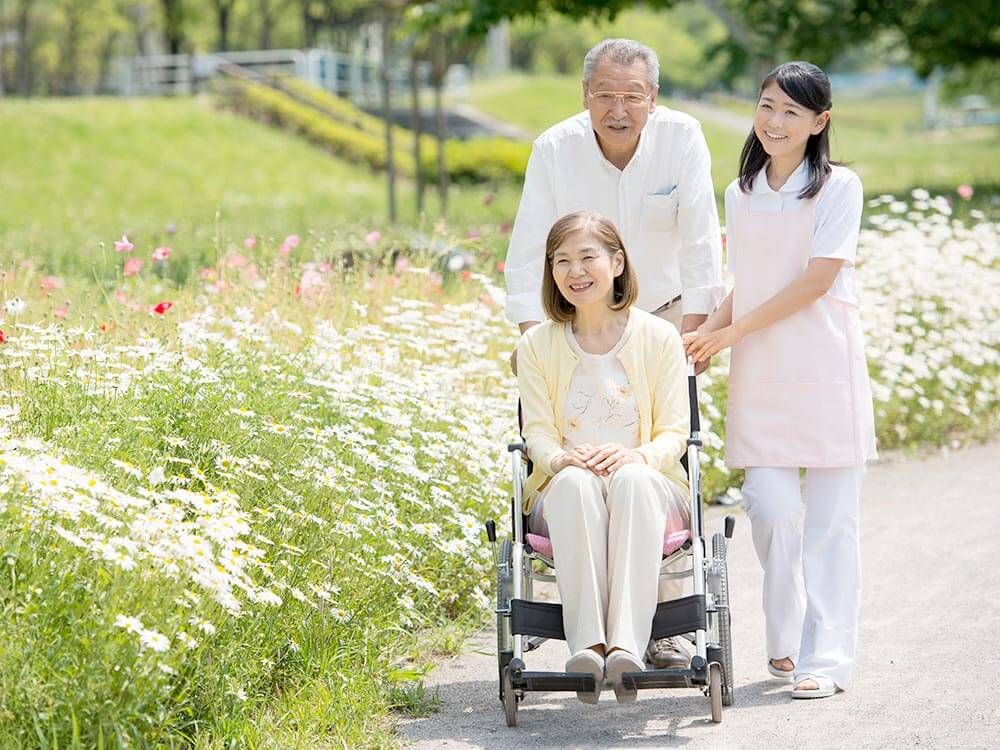 花の咲く道を散歩する車椅子に座っている高齢女性と介助をするヘルパー