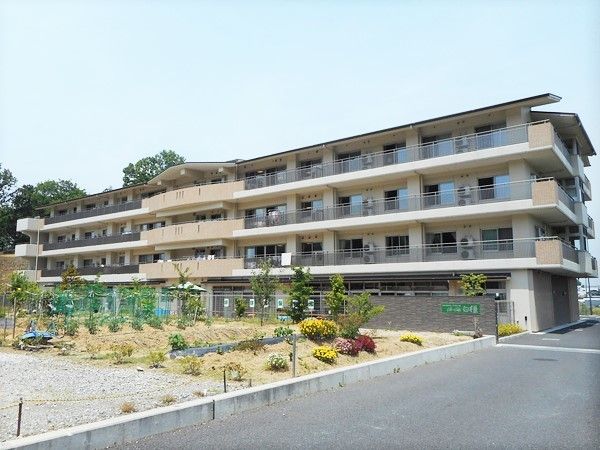 ４階建てのブラウン系の建物は横に長い長方形で重厚感があり、広い野菜畑が手前にある。