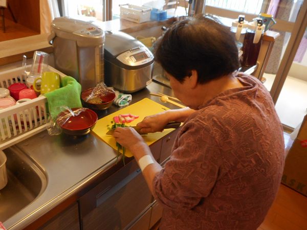 黄色いまな板の上で野菜を切っている女性入居者。キッチンには炊飯器や電気ポットが置かれている。