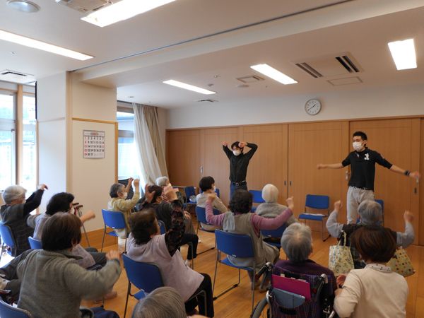 体操に励む入居者と指導に当たるスタッフの様子。ホールに集まった入居者がいすに座って体を動かしている。