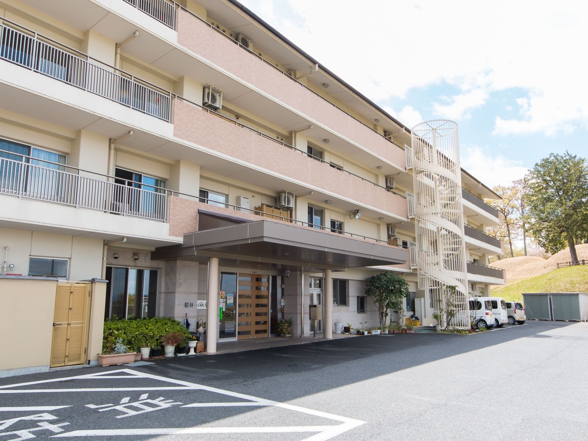 ４階建ての施設である。玄関前には大きなひさしがついている。建物の前には広いスペースがあり、車が停められている。