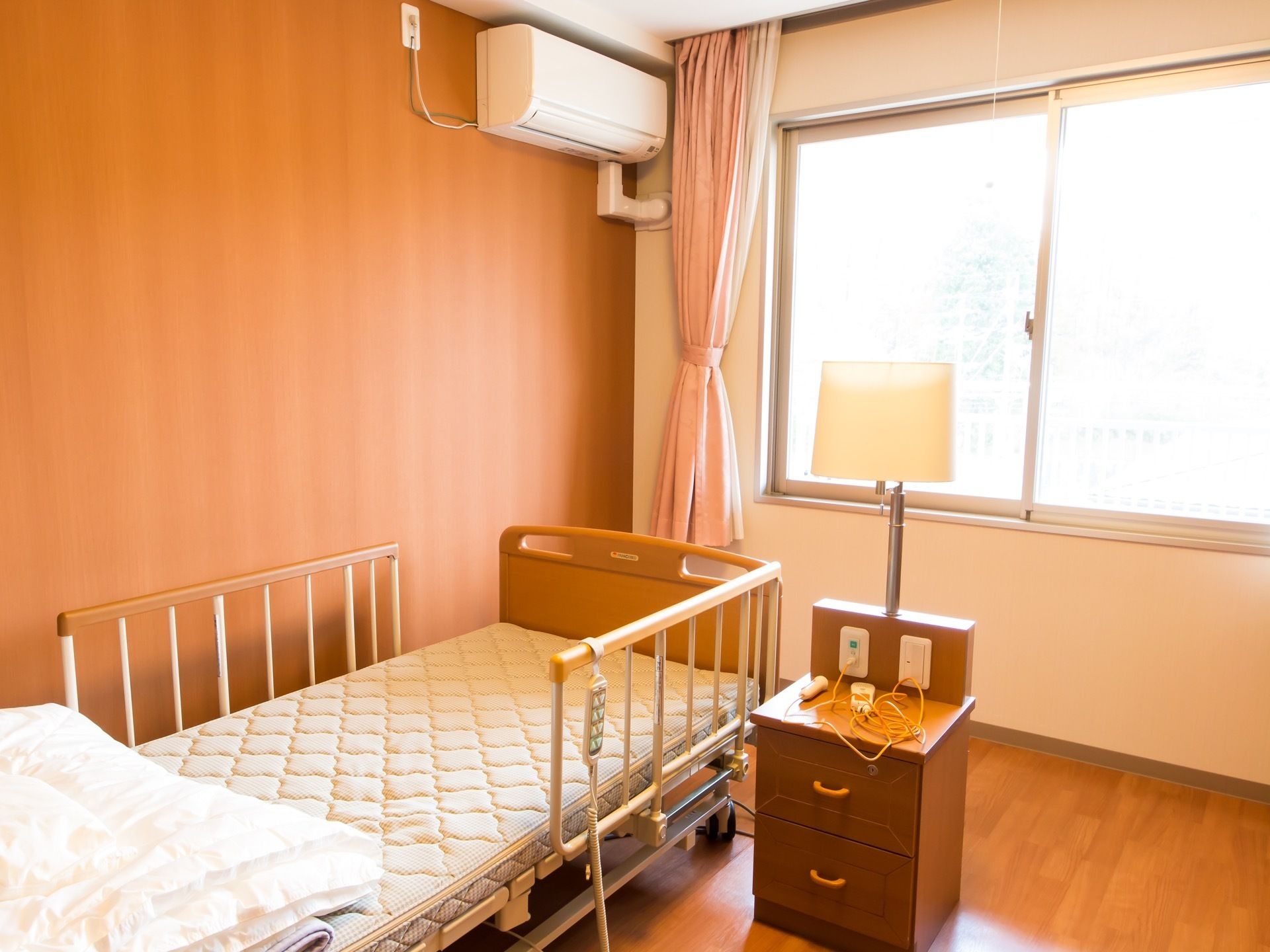 居室には大きな窓がある。転落防止柵のついた介護ベッドやエアコンが設置されている。電気スタンド付のサイドボードが置かれている。