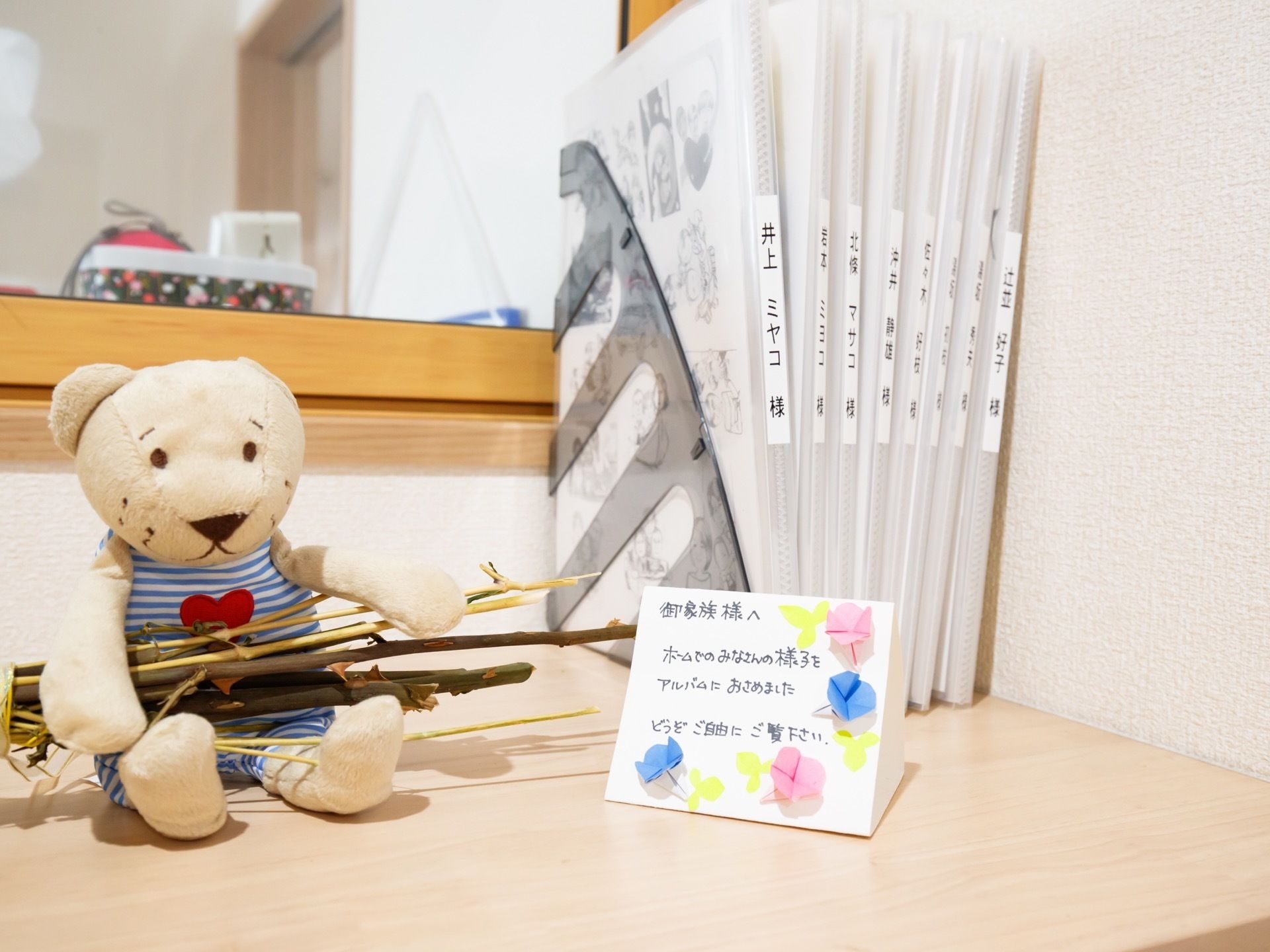 カウンターの上にクマのぬいぐるみと8冊ほどの写真ファイルが置かれている。そばにある紙には、入居者様のご家族あてのメッセージが書かれている。