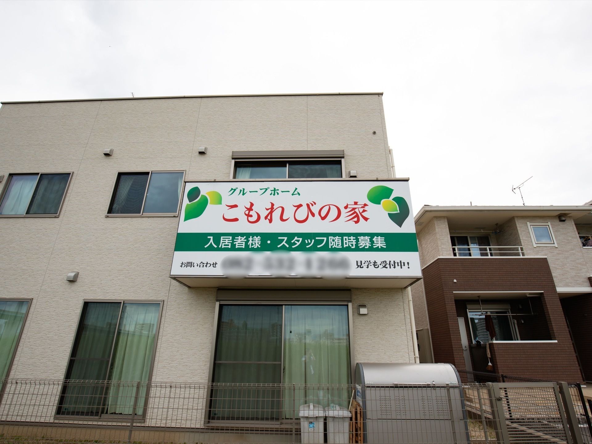 施設は2階建てで、窓にはカーテンがかけられている。施設の大きな看板があり、施設名と入居者様やスタッフ募集の旨が書かれている。