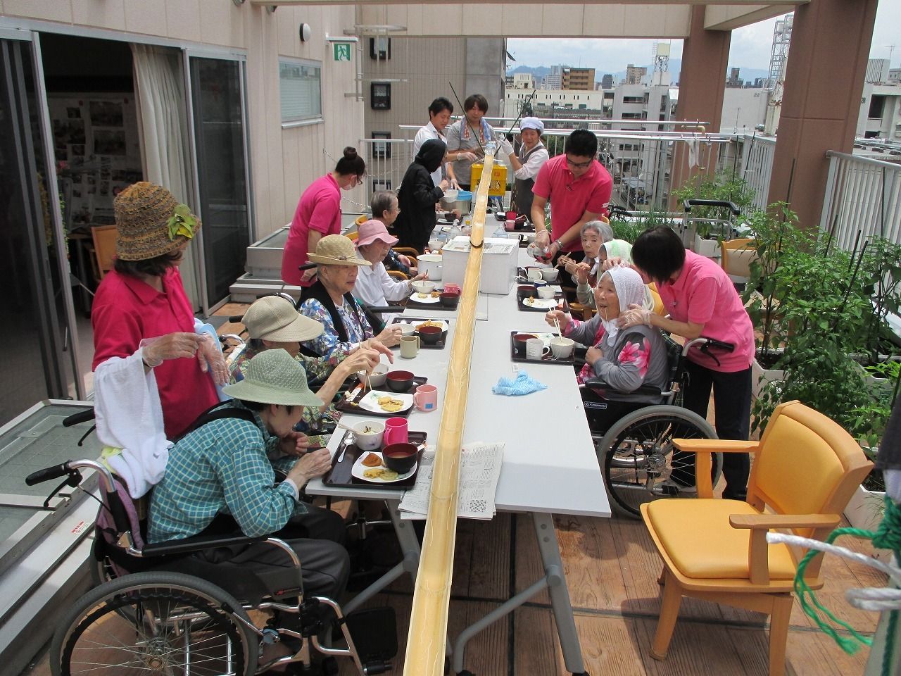 ホームの外に机を並べ、帽子をかぶってそうめん流しを楽しむ入居者と、それを周りで見守るスタッフ。