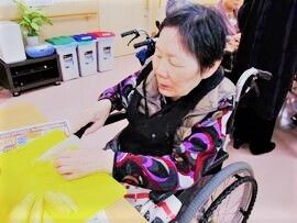 車いすの女性入居者が包丁を持って野菜を切っている様子。周りには分別用のゴミ箱や観葉植物が置かれている。