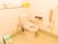 ベージュの床と白い壁の広いトイレがある。壁には手すりが設置されており、棚にはトイレ用品が置かれている。
