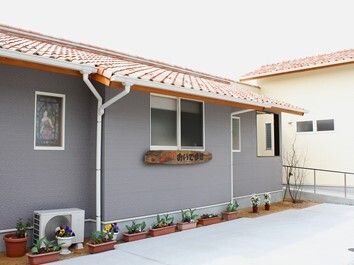 平屋建ての建物はグレーで、外壁に木製の看板があり、ホーム名が書かれている。建物の周りには花が植えられたプランターが置かれている。