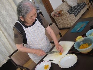 エプロンを付けた白髪の女性が真剣な表情で野菜を切っている様子。白いまな板は野菜の色が染みてオレンジのようになっている。