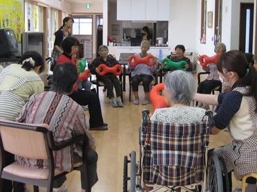 円になって椅子に座り、３B体操をしている入居者たち。そばにはエプロンを付けた女性スタッフたちがいて、入居者は３B体操の道具を持っている。