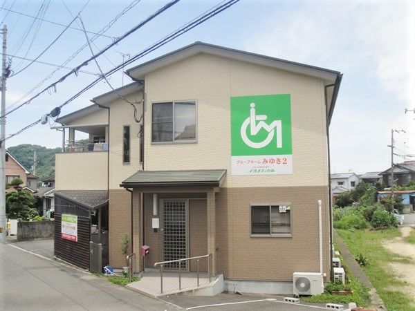 大きめの一軒家風のデザインの建物。奥のほうに住宅街と自然豊かな場所がある