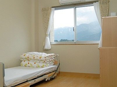 レースカーテンとカーテンが付いた大きな窓があるフローリングの部屋には寝具付きのベッドやエアコンがある。