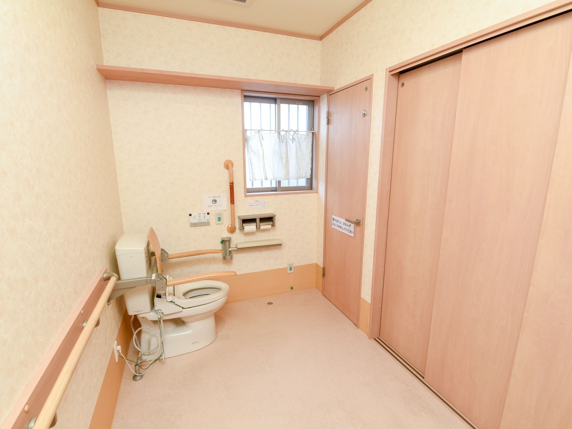 ベージュの壁に囲まれた広いトイレがある。窓がついており、壁には手すりがある。便座には背もたれとひじ掛けが設置されている。