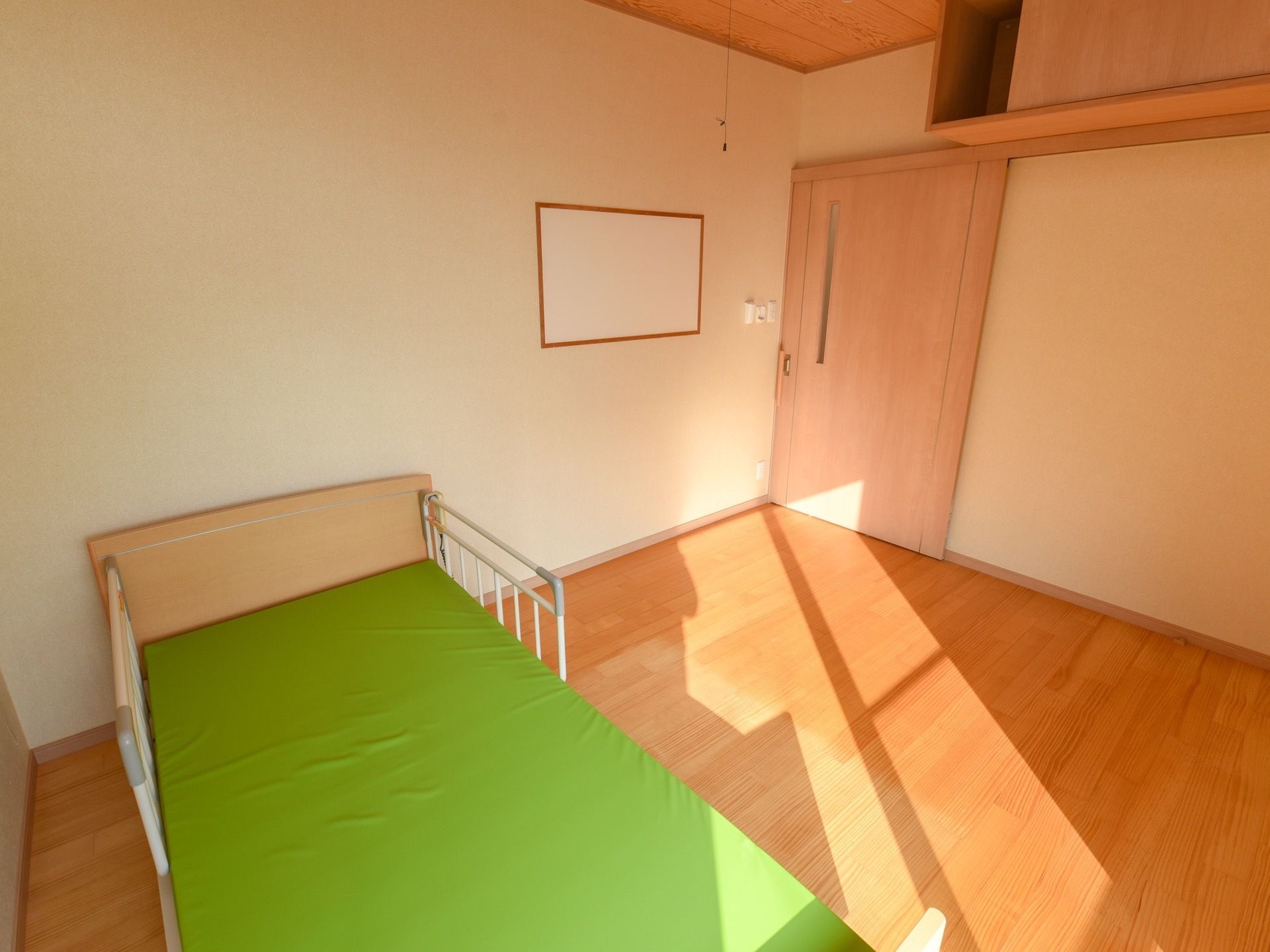 木目調の床の、日当たりの良い居室がある。介護用ベッドが置かれている。天井付近には棚が設置されている。