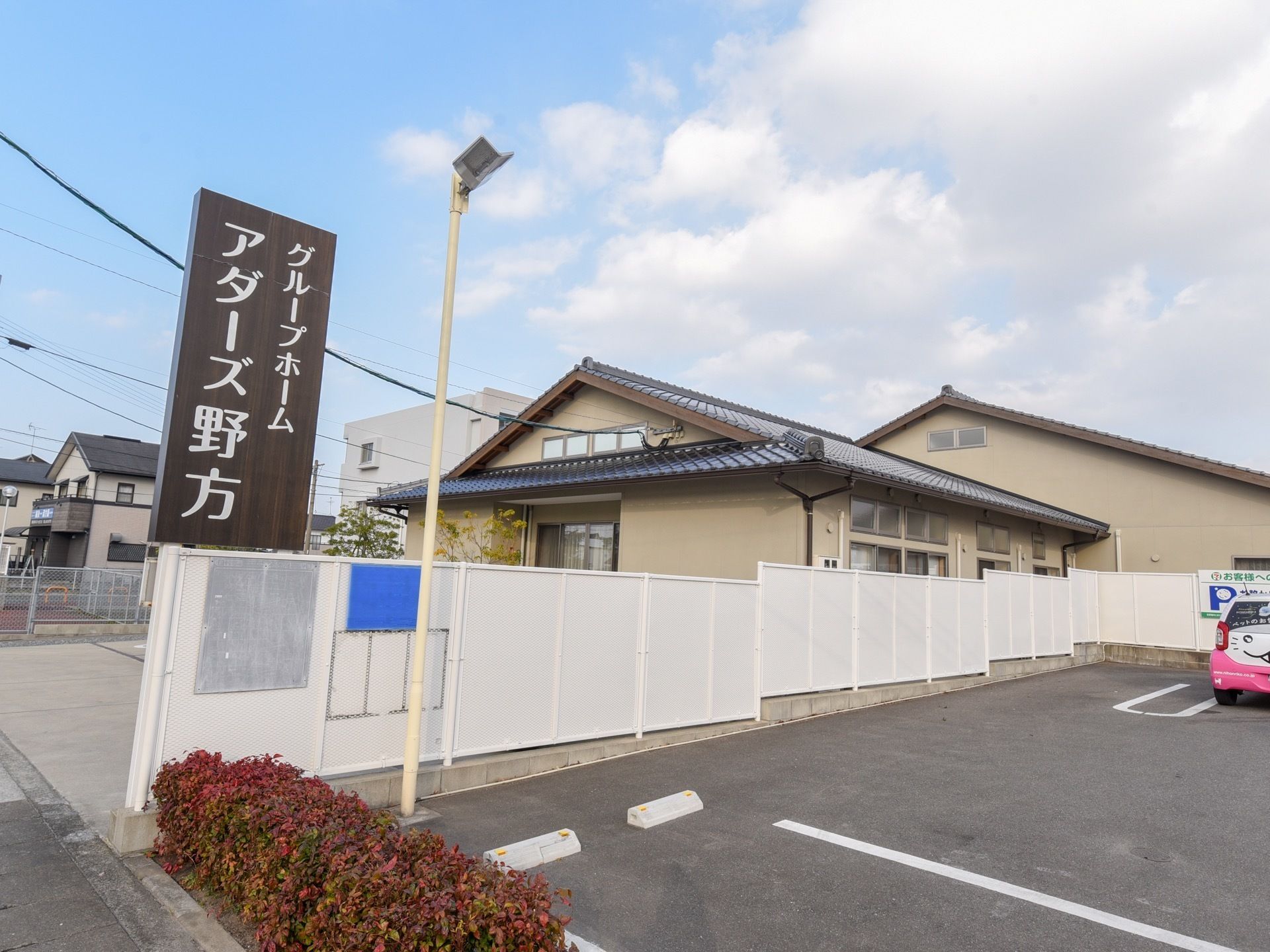 グレーの壁と瓦屋根の日本家屋風の建物がある。敷地の周りは白いフェンスで囲まれている。入り口には施設名が書かれた看板がある。