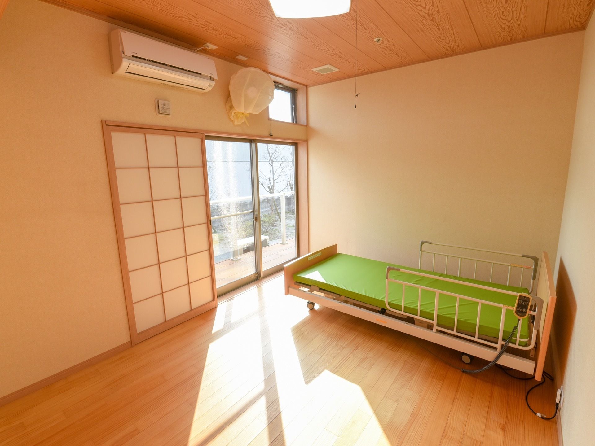 木目調の天井と床の広い居室がある。窓には障子がはまっている。介護用ベッドが置かれ、緑色のマットレスが敷かれている。