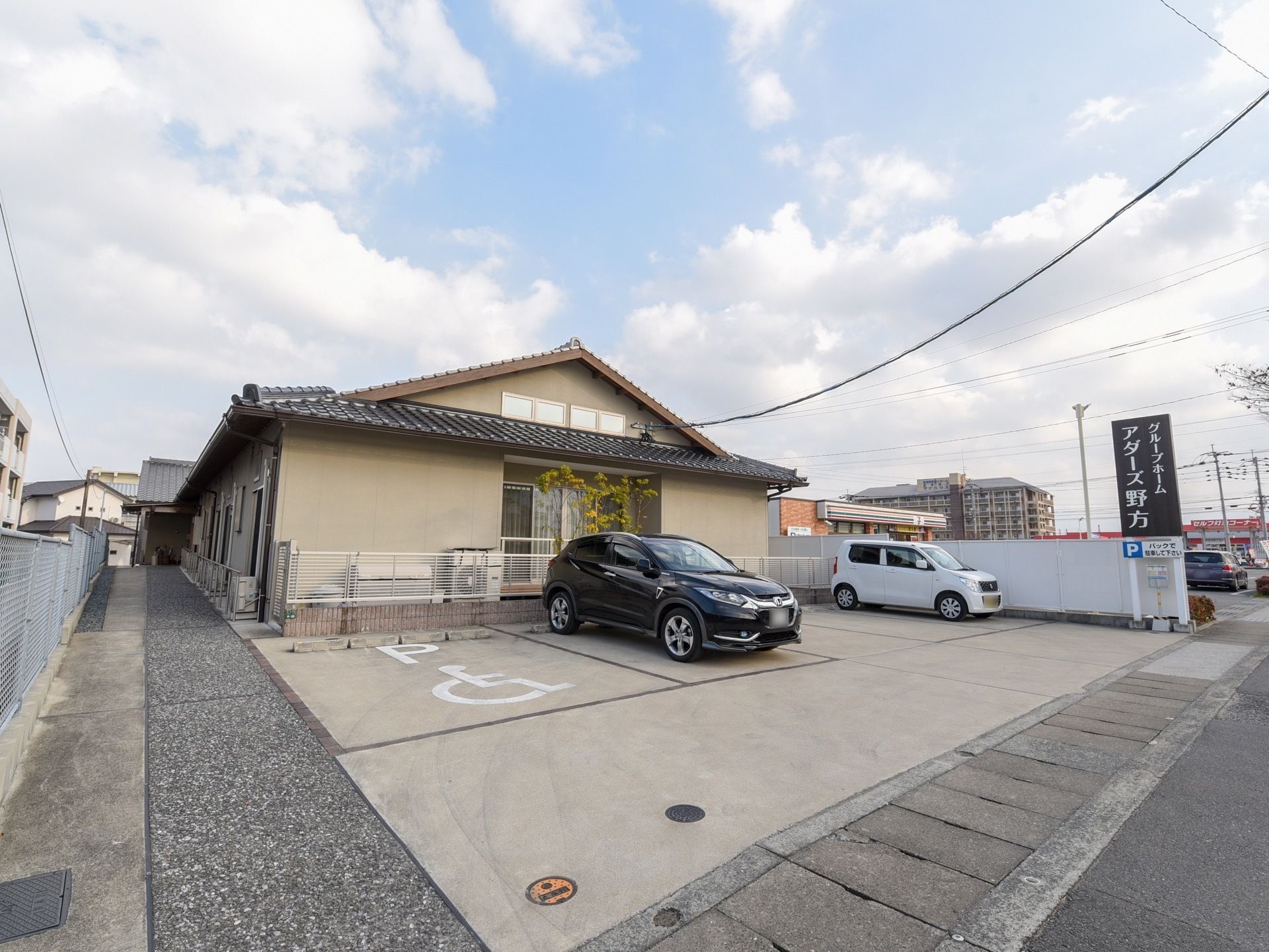 日本家屋風の平屋建ての建物がある。周りは白いフェンスで囲まれ、道路沿いが広い駐車場になっており、車が停まっている。